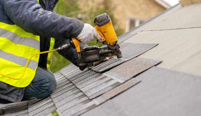 A close-up of a roofer wearing a safety vest and gloves, using a pneumatic nail gun to install grey asphalt shingles on a roof.
